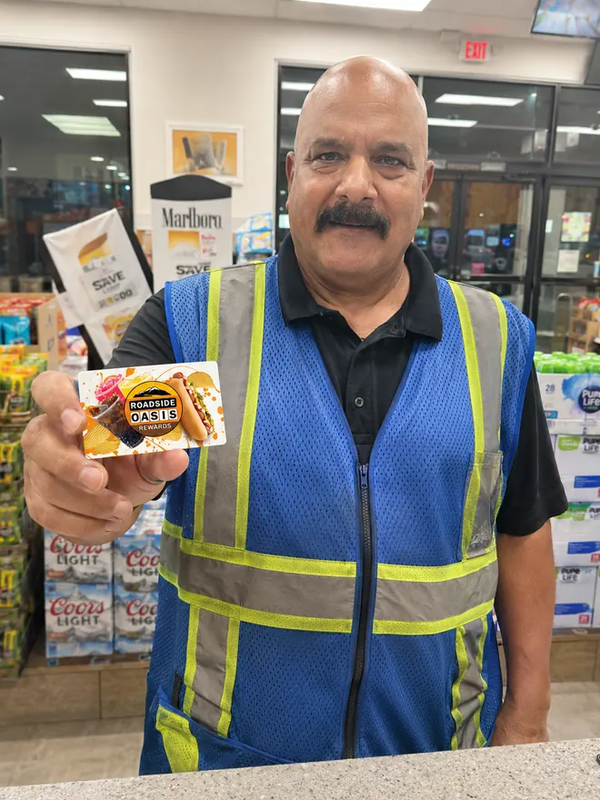 Roadside Oasis employee in a blue hi-vis vest holding a Rewards card inside the convenience store in Menifee, CA