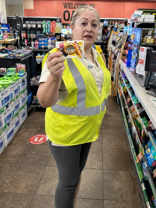 Store associate in a yellow hi-vis vest holding a Roadside Oasis Rewards card in the snack aisle in Menifee, CA