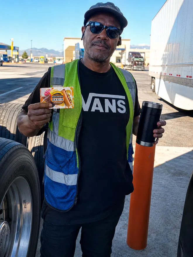 Truck driver in sunglasses and a safety vest holding his Rewards card beside his trailer at the fuel island in Menifee, CA